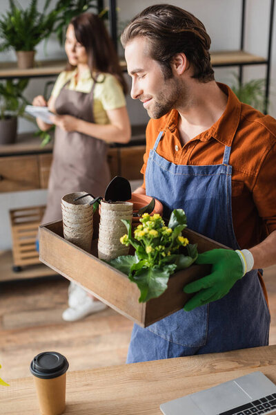 Young florist holding box with gardening tools and flowerpots near laptop and coffee in flower shop 