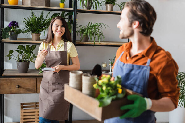 Smiling florist in apron holding notebook near blurred colleague with box in flower shop 