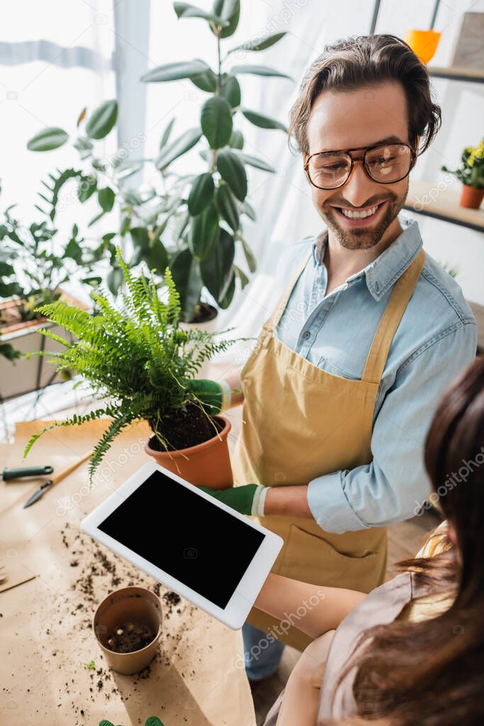Positive florist holding plant and flowerpot near colleague with digital tablet in flower shop 