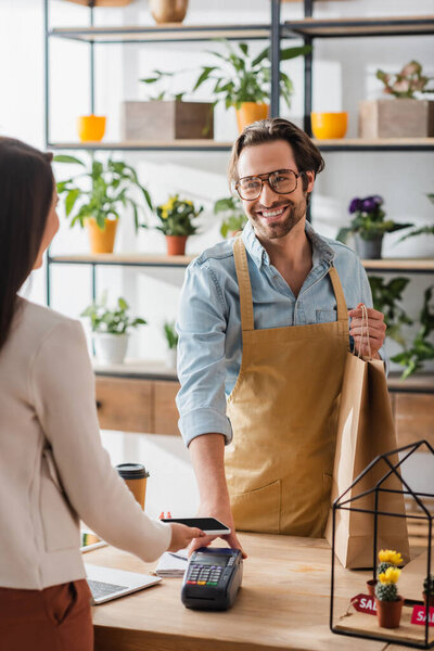 Smiling florist holding shopping bag near payment terminal and blurred customer with smartphone in flower shop 