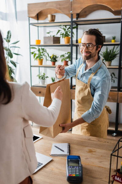 Smiling florist holding shopping bag near blurred customer in flower shop 