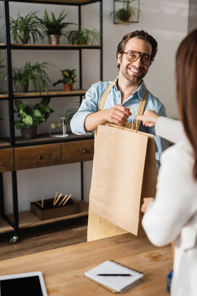 Happy florist holding shopping bag near blurred customer and digital tablet in flower shop 