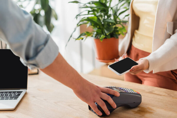 Cropped view of florist holding payment terminal near customer with smartphone in flower shop 