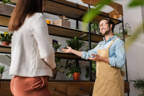 Low angle view of smiling florist pointing at plants near blurred customer in flower shop 