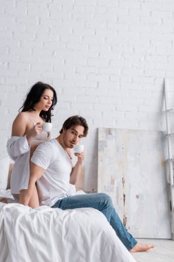 Kot pantolonlu adam yatak odasında beyaz gömlekli ve sütyenli seksi bir kadının yanında kahve içiyor.