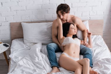 Kot pantolonlu üstsüz bir adam ve beyaz yatağa sarılmış tutkulu bir kadın.