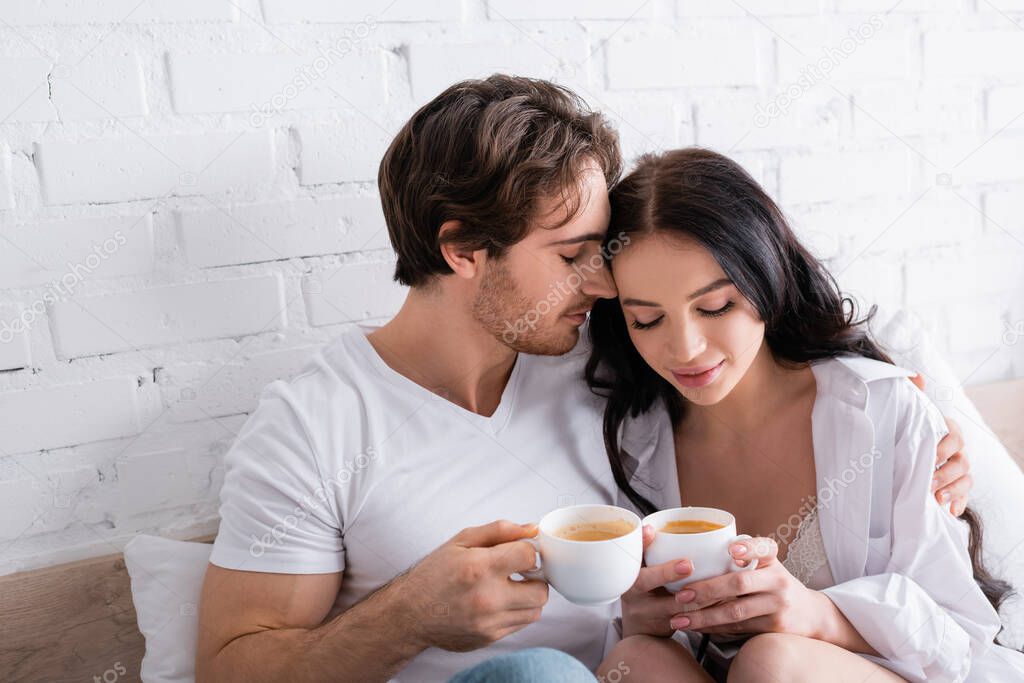 happy young couple sitting in bed with cups of coffee and embracing with closed eyes