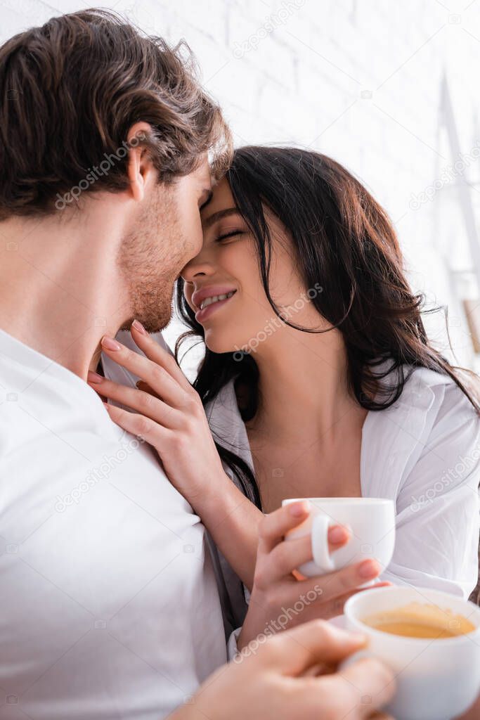 sensual brunette woman and young man holding coffee while embracing in bedroom