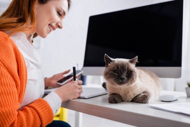 Akıllı telefonlu gülümseyen kadın masadaki kedinin yanında deftere yazı yazıyor.