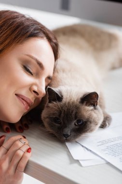 Mutlu bir kadın, gözleri kapalı, masasında tüy gibi bir kedinin yanında gülümsüyor.
