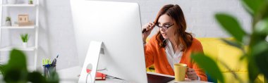 woman with cup of tea touching eyeglasses while working near monitor on blurred foreground, banner clipart