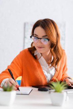 Gözlüklü gülümseyen kadın evdeki grafik tabletinin yanında deftere yazıyor.