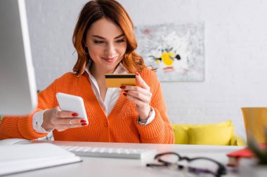 Evde çalışırken elinde kredi kartı ve akıllı telefon tutan gülümseyen bir kadın.