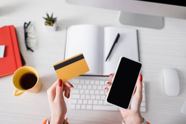 Bulanık defter, klavye ve çayın yanında elinde kredi kartı ve akıllı telefon tutan bir kadının üst görüntüsü.