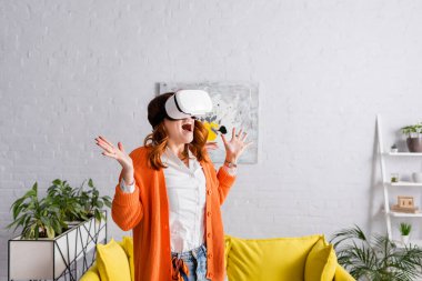 VR kulaklıklı hayretler içinde bir kadın oturma odasında oyun oynarken yaptığı hareketi gösteriyor.