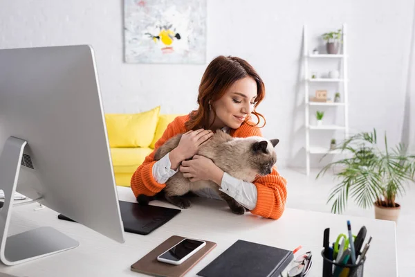 happy freelancer holding cat while sitting at work desk near graphic tablet and computer