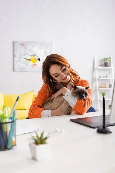 happy woman embracing cat while sitting at desk near blurred graphic tablet and stylus