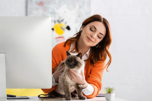 smiling woman cuddling cat on work desk near computer monitor