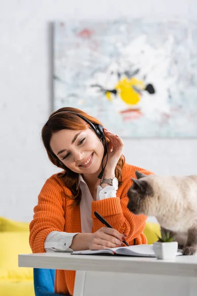 Cheerful Woman Headset Writing Notebook While Working Cat Sitting Desk — Stock Photo, Image