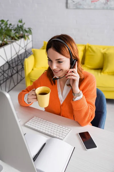 Mujer Sonriente Auriculares Sosteniendo Taza Mientras Trabaja Cerca Computadora Borrosa — Foto de Stock