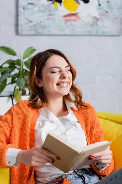 本とソファに座っている間に目を閉じて笑う陽気な女性 — ストック写真