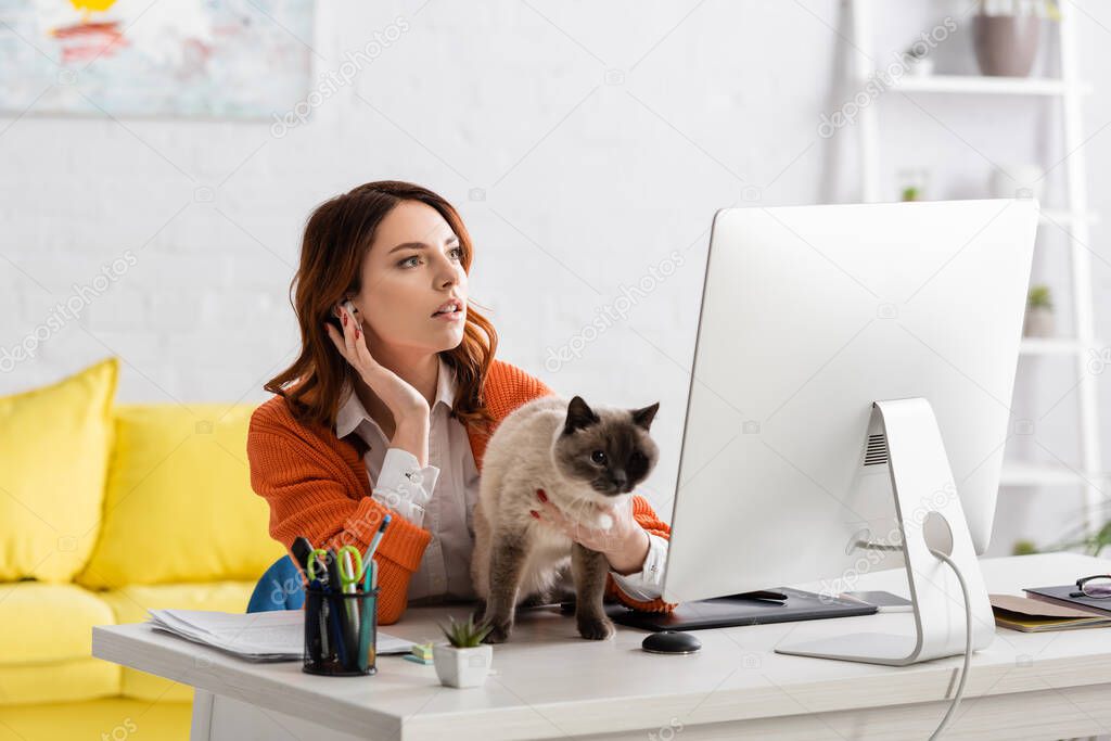 young designer inserting earphone while sitting near computer monitor with cat