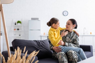 Smiling woman in camouflage uniform hugging daughter on couch at home  clipart