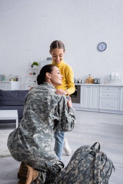 Gülümseyen bir kadın, evde askeri çantanın yanında çocuğa sarılıyor. 