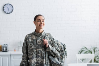 Cheerful woman in military uniform holding backpack at home  clipart