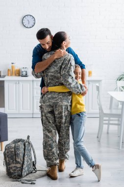 Husband and daughter hugging mother in military uniform near backpack at home  clipart