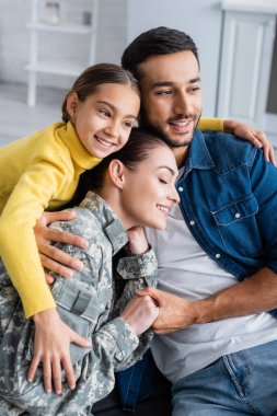 Positive woman in military uniform holding hand of husband near kid at home  clipart