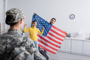 Çocuğun yanında Amerikan bayrağı tutan mutlu adam evet işareti yapıyor ve evde askeri üniformayla annesini bulanıklaştırıyor. 