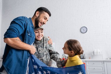 Asker üniformalı bir adam, mutfakta Amerikan bayrağında kızının yanında karısına sarılıyor. 