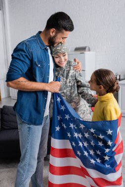 Amerikan bayrağı taşıyan neşeli bir adam ve evde kızının yanında kamuflaj üniformalı bir eş. 