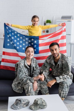 Mutlu çocuk Amerikan bayrağını elinde tutuyor askeri üniformalı ebeveynlerin yanında evdeki kanepede. 