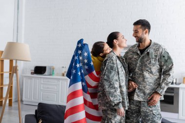 Amerikan bayrağını elinde tutan çocuk evde askeri üniformalı gülümseyen ebeveynlerin yanında. 
