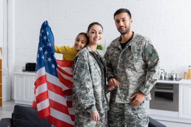 Parents in military uniform holding hands near preteen kid with american flag at home  clipart