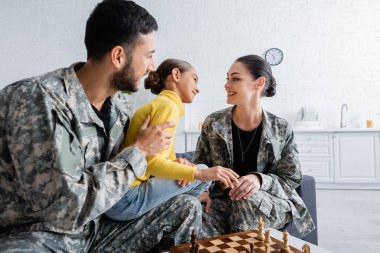 Askeri üniformalı pozitif ebeveynler kızlarının yanında oturuyor ve evde satranç oynuyorlar. 