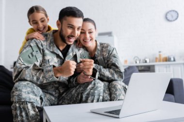 Laptop near man in military uniform showing yes gesture near wife and daughter on blurred background  clipart