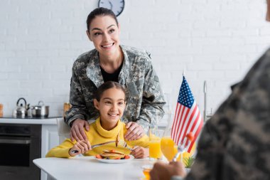 Mutfakta kahvaltıda kızının ve Amerikan bayrağının yanında duran kamuflaj üniformalı gülümseyen anne. 