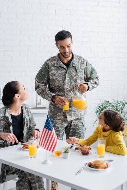 Man in military uniform pouring orange juice near family and american flag during breakfast at home  clipart