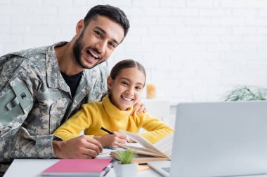 Askeri üniformalı neşeli bir adam evde dizüstü bilgisayar ve defterlerin yanında kızına sarılıyor. 
