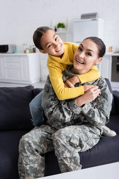 Positive girl embracing mom in military uniform on couch at home