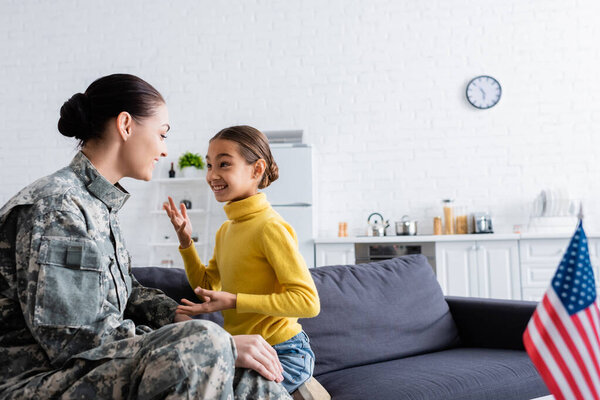 Smiling kid talking to mother in military uniform near blurred american flag at home 