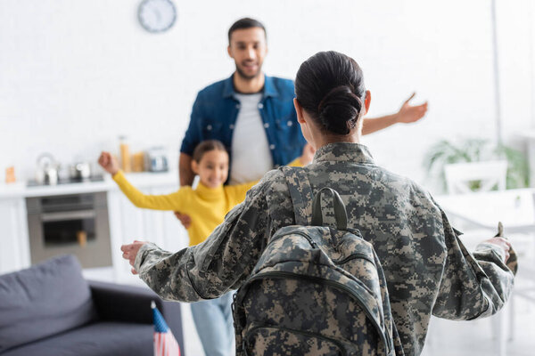 Woman in military uniform standing near blurred husband and daughter at home 