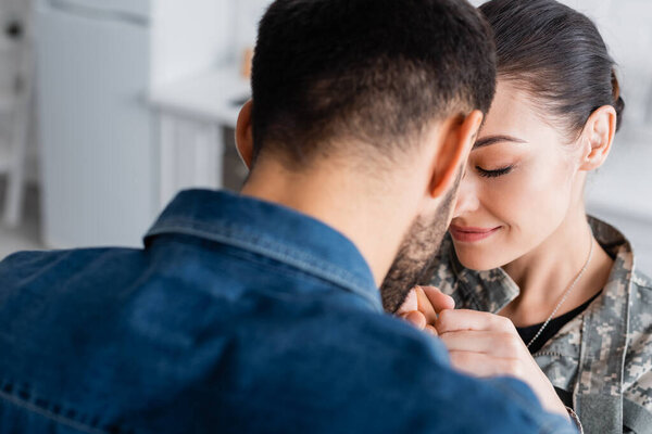 Blurred man holding hands of wife in military uniform at home 