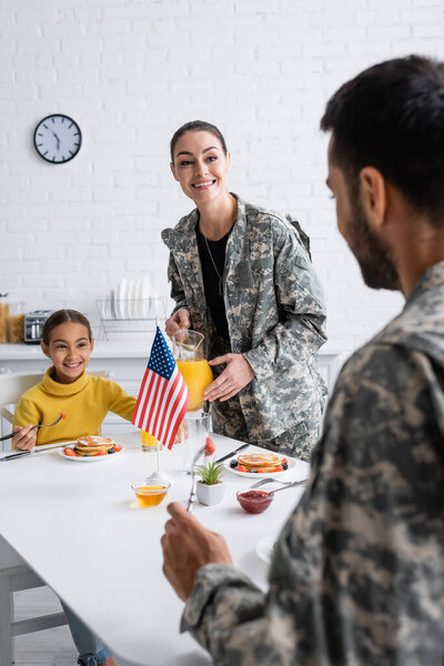 Smiling woman in military uniform holding orange juice near daughter and husband during breakfast at home 