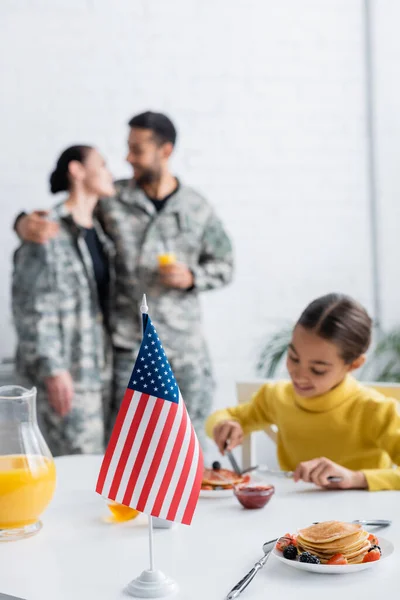 带早餐的孩子和背景模糊的身着军服的父母身边的美国国旗 — 图库照片