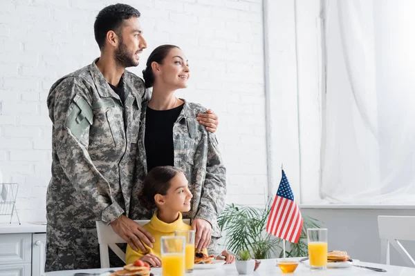 家で朝食中に娘とアメリカの旗の近くに立ってカモフラージュで陽気な両親 — ストック写真