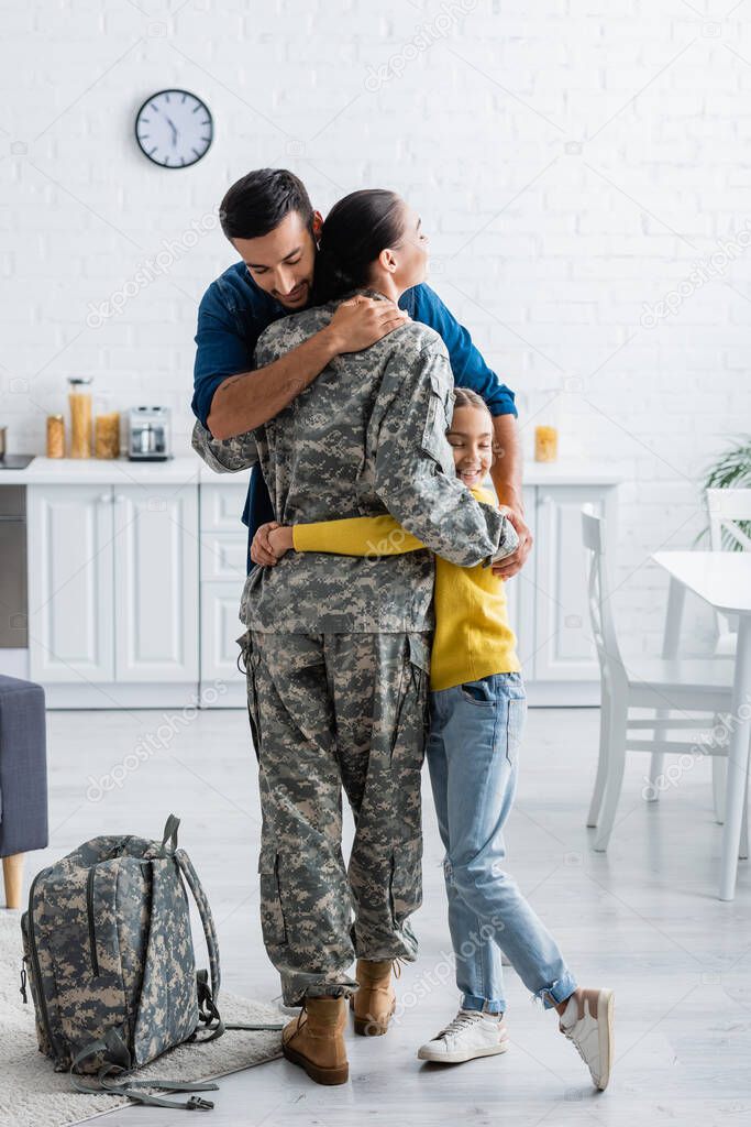 Husband and daughter hugging mother in military uniform near backpack at home 